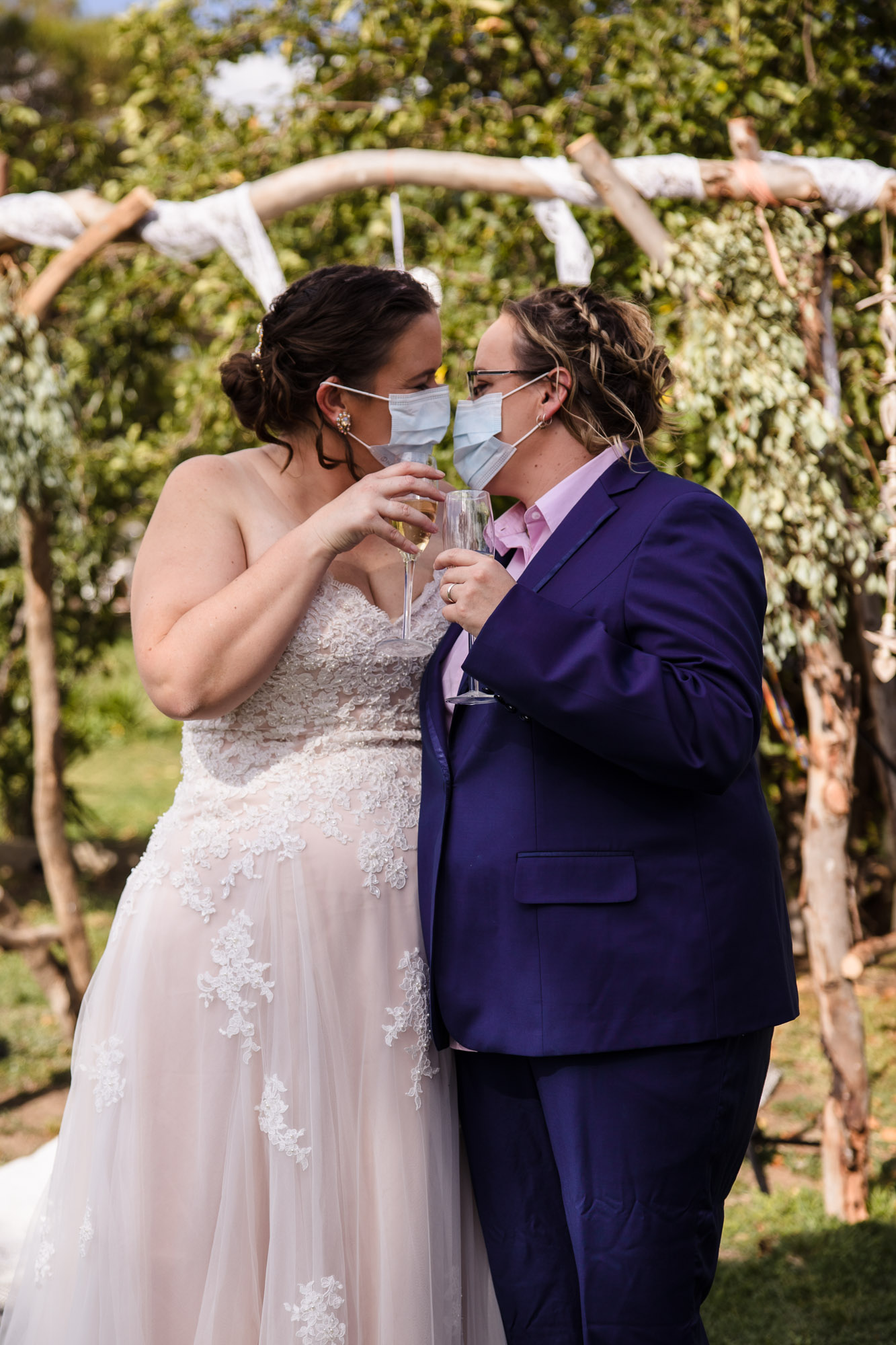 two masked brides kiss with glasses of champagne in hands at covid 19 wedding elopement 